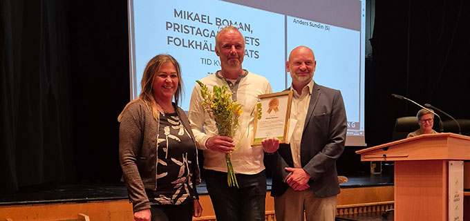 En bild på en kvinna och två män som står på en scen. Mannen i mitten håller i ett diplom och en blomma.