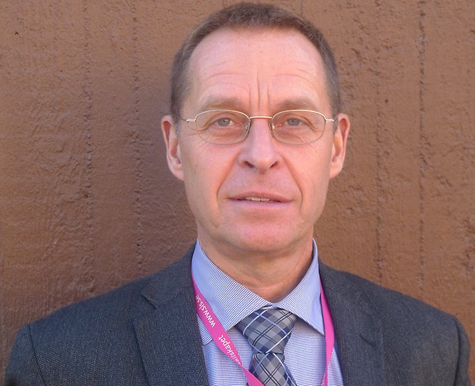 Huvudbild på en person i kostym och glasögon som står mot en brun yttervägg