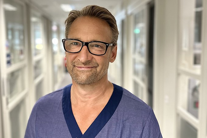Halvkroppsbild på person i blå läkarkläder med glasögon och kort brunt hår som ler in i kameran.
