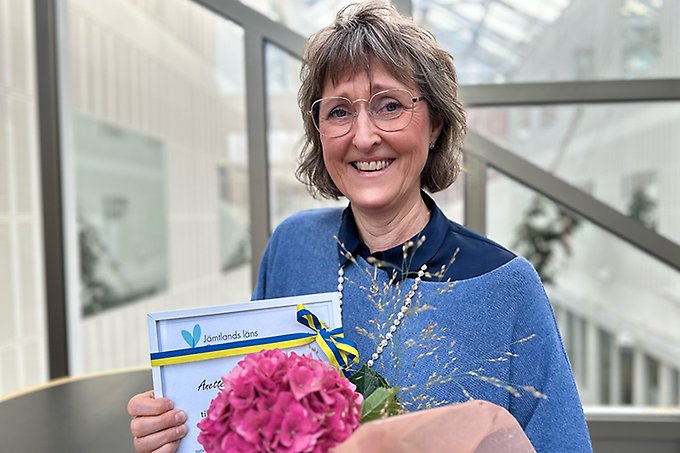 Person står vid ett runt bord med blommor och ett stipendium i händerna. Ler mot kameran.