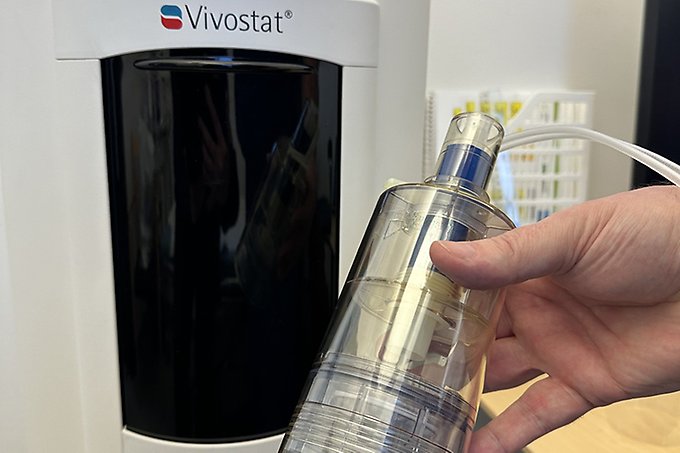 Närbild på en hand som håller i en plastbehållare intill en medicinteknisk apparat.
